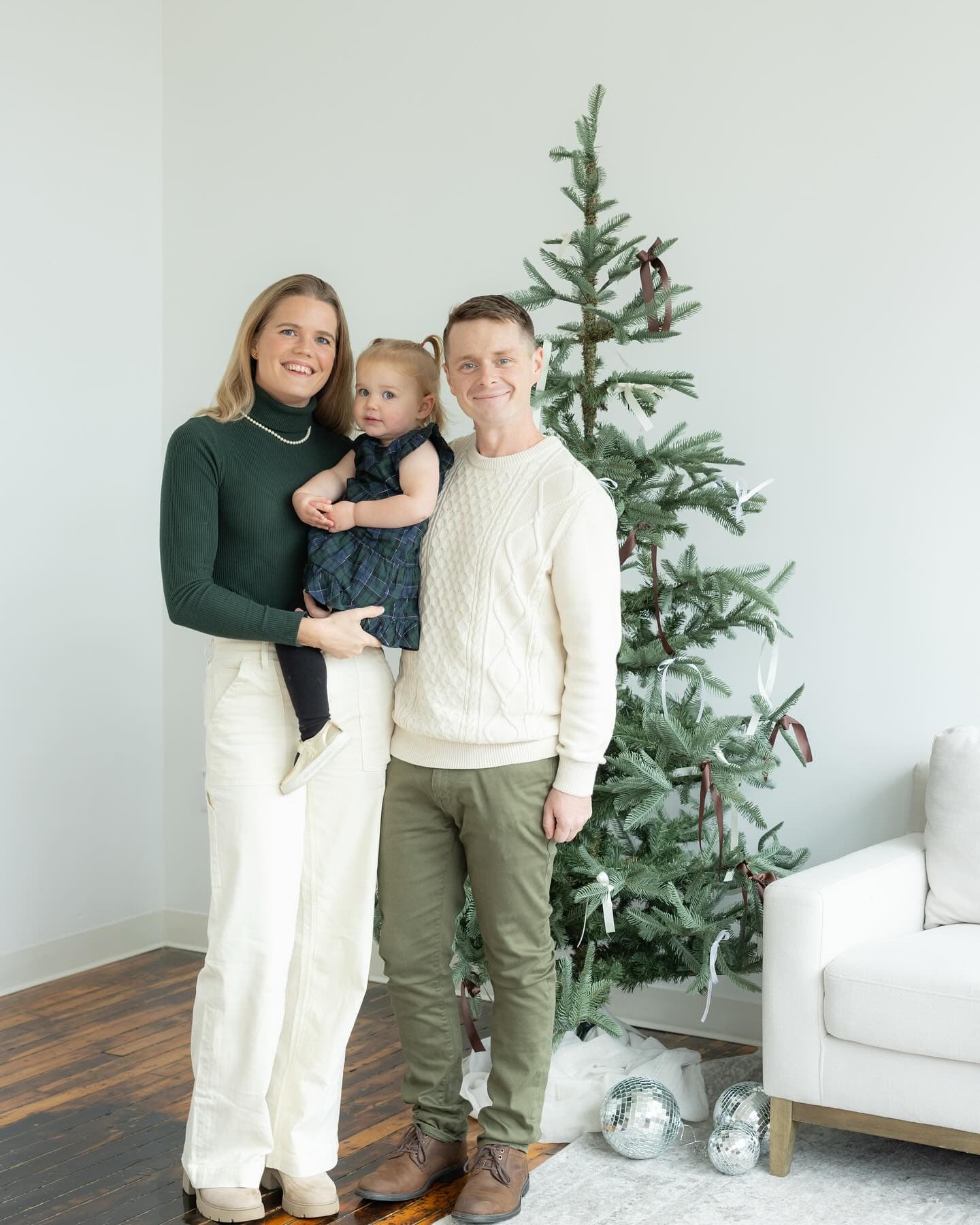 A family of three stands together in front of a Christmas tree, smiling for the camera.