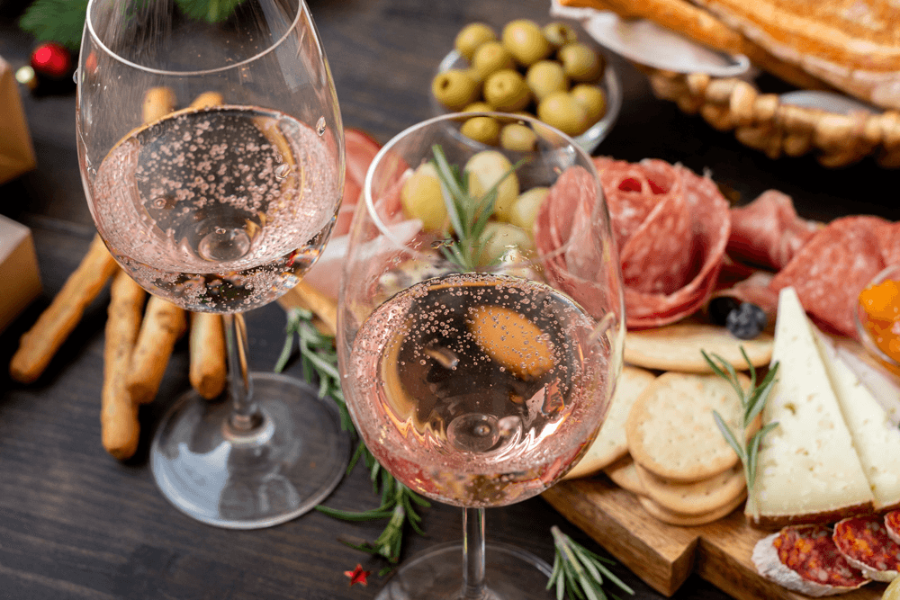 Two glasses of sparkling rosé wine sit amidst a spread of olives, meats, cheeses, and breadsticks on a wooden table.