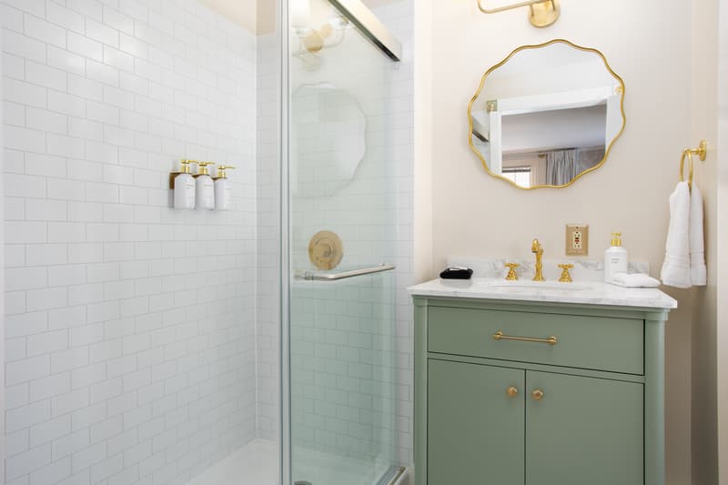 A modern bathroom featuring a glass shower, mint green vanity, and gold fixtures.