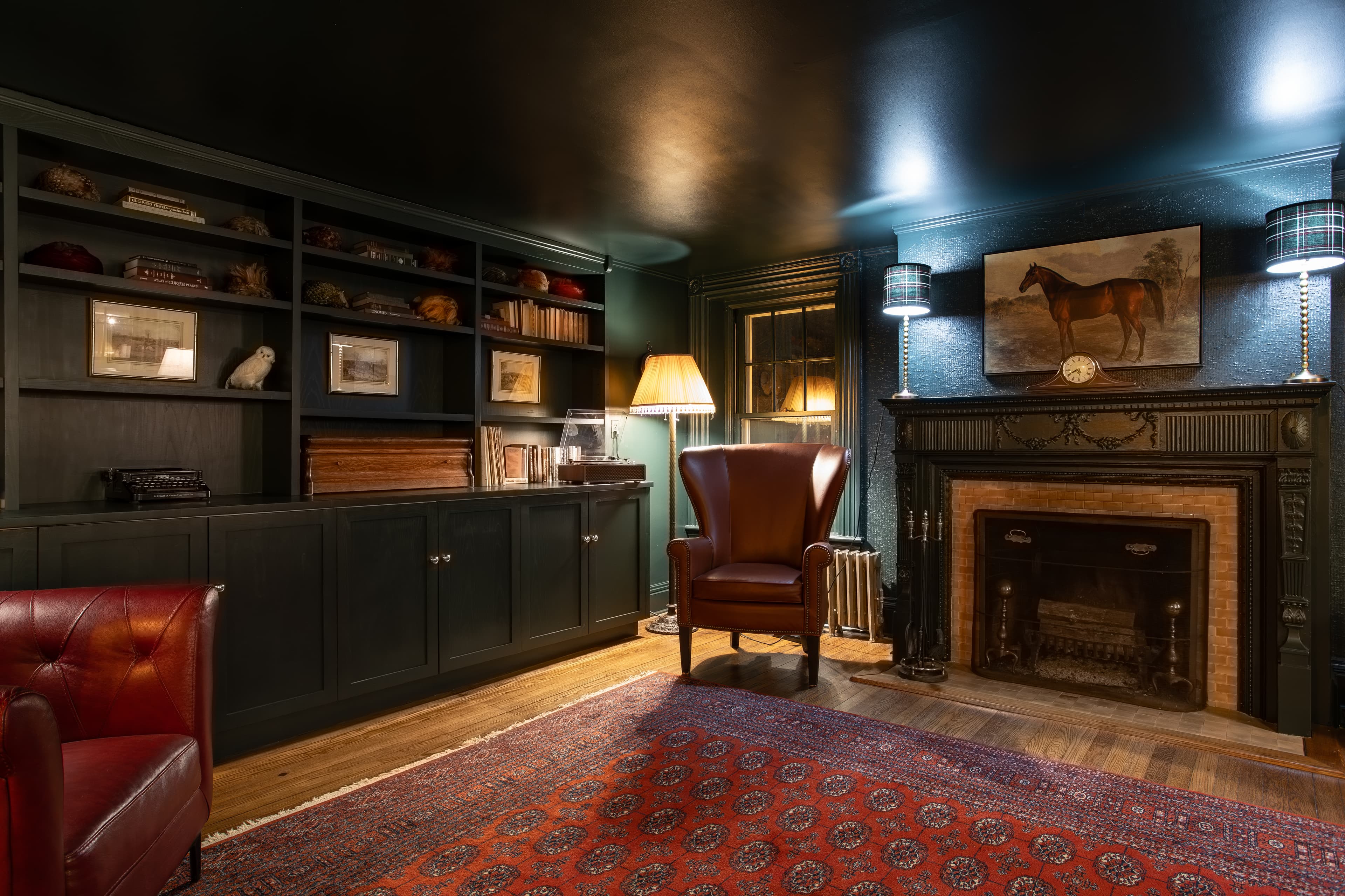 A cozy, dimly lit room featuring a leather armchair, dark shelves filled with books, and a fireplace adorned with a horse painting.