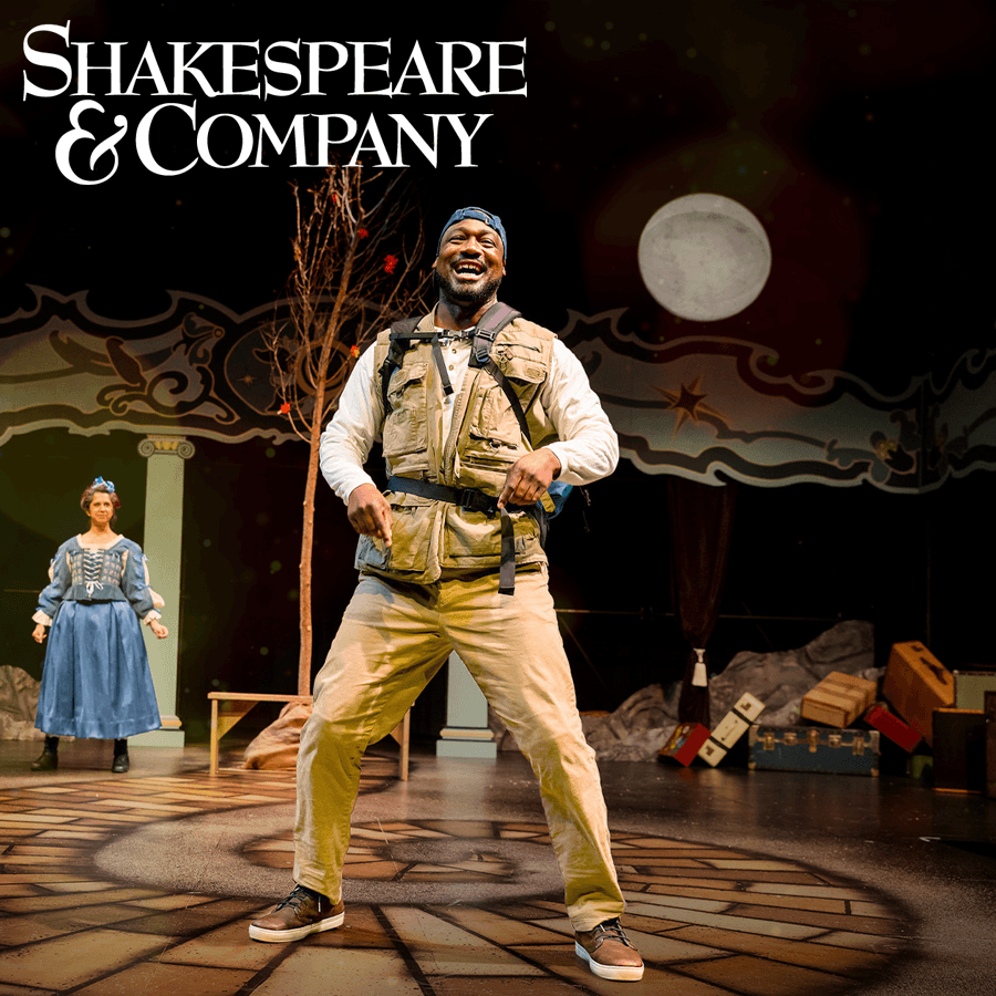 A smiling performer in casual clothing stands on stage, with a woman in a blue costume in the background.