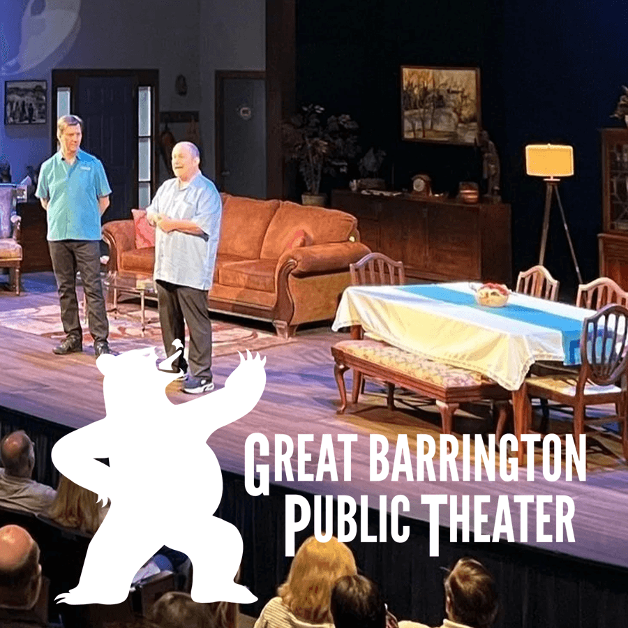 Two actors perform on stage in a theater setting, with an audience visible in the foreground.