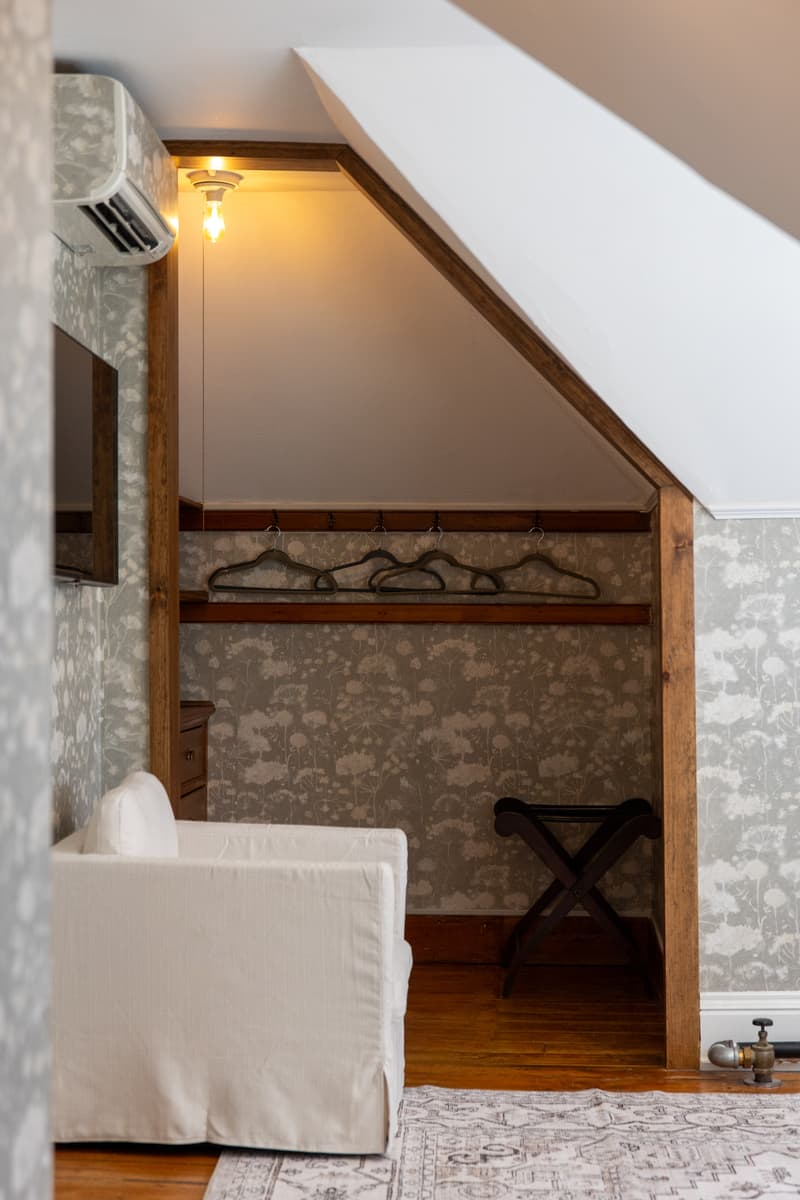 A cozy attic corner with a white sofa, a wooden hanger rack, and soft lighting against floral wallpaper.